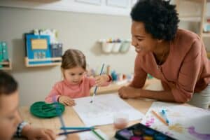 Happy black teacher assisting small girl in drawing during art class at kindergarten.